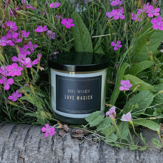 A candle surrounded by herbals with a label that reads 'Love Magick' This candle is designed to invoke feelings of love and attraction with its sweet, seductive scents. Crafted to attract new love, promote self-adoration, and foster harmonious relationships. Made with soy blend wax and cotton core wicks, free from unsafe synthetic fragrances. Offers a clean burn and minimal soot when properly trimmed.